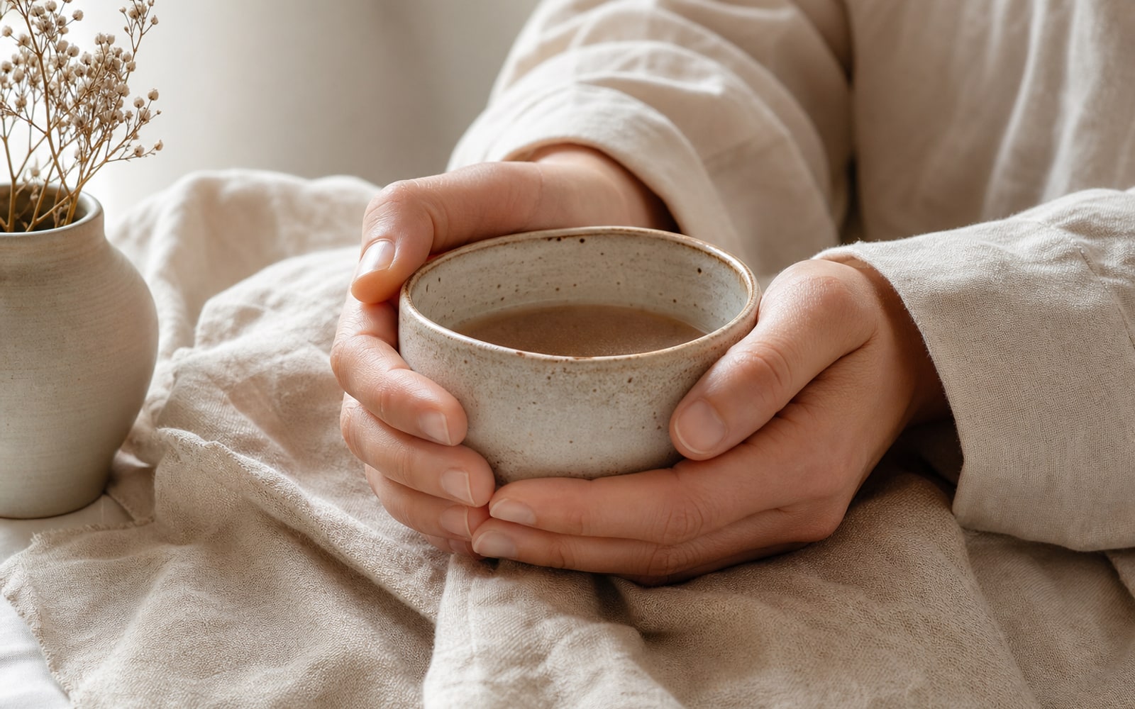 Hands holding a warm ceramic cup over linen in soft daylight