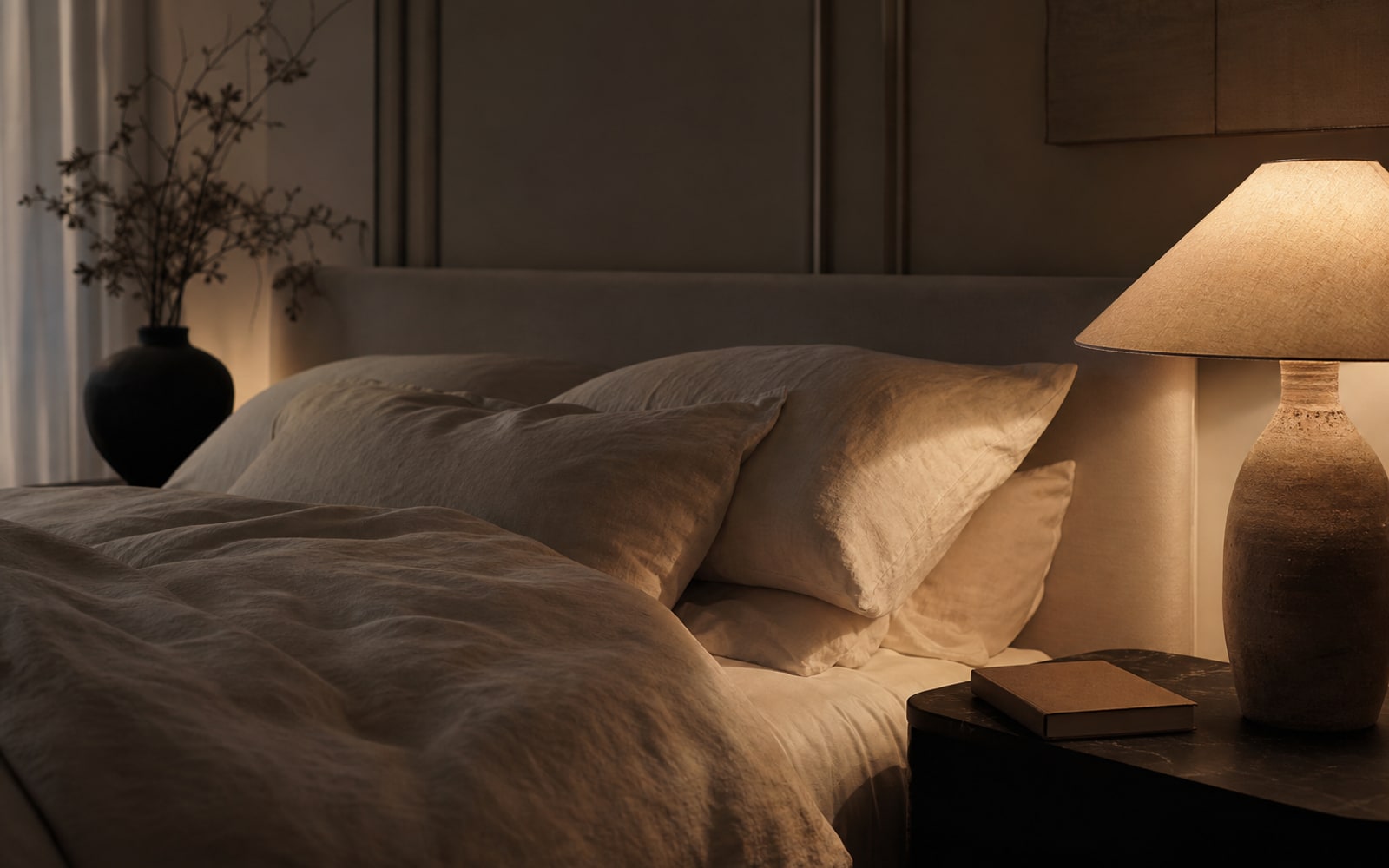 Dim bedroom at night with linen bedding and a closed book by soft lamplight